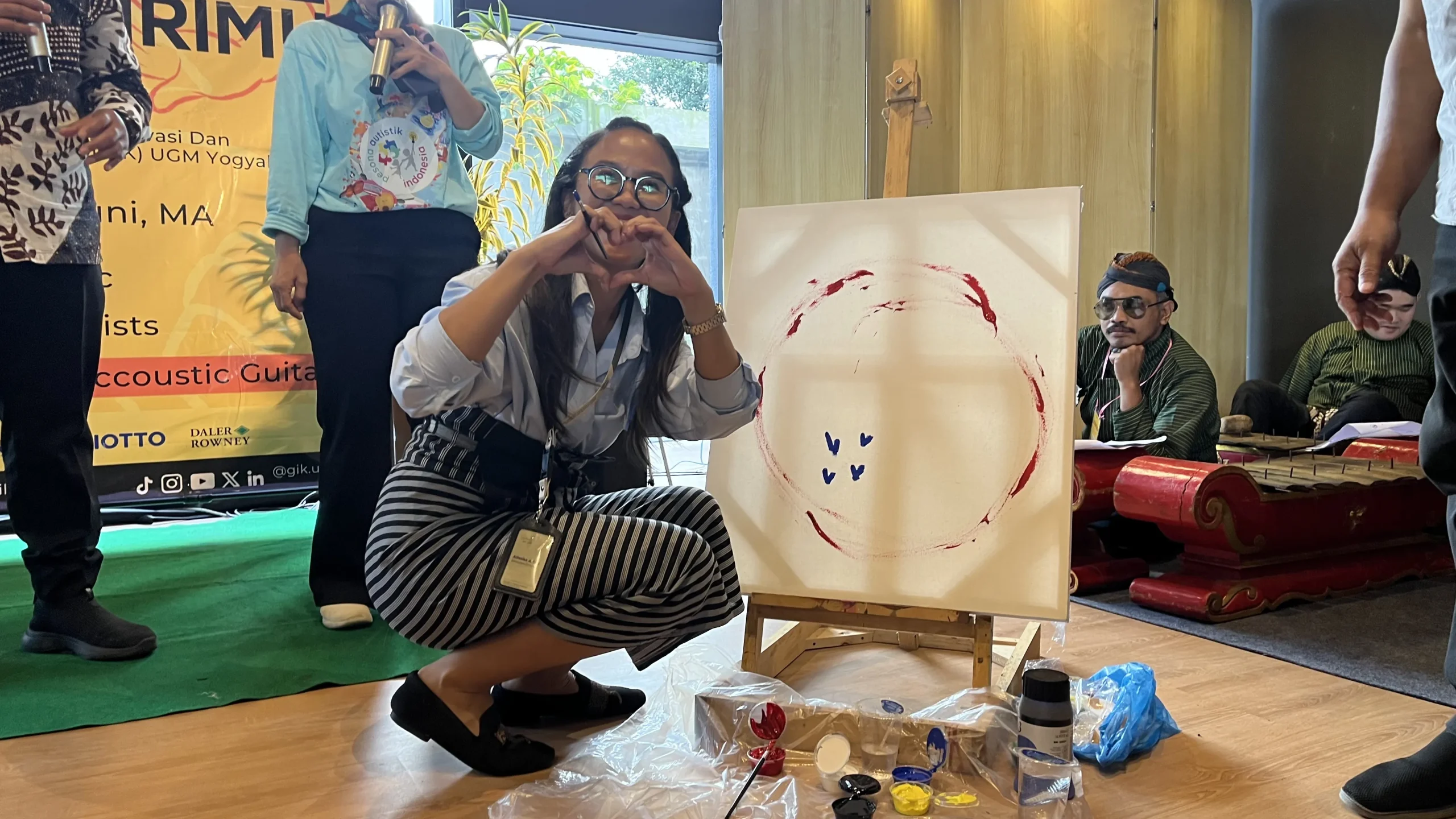 Bu Alfatika (Direktur GIK UGM) tersenyum dan membentuk simbol hati dengan tangan di depan kanvas lukisan saat sesi live painting dalam rangkaian acara IPED di GIK UGM.