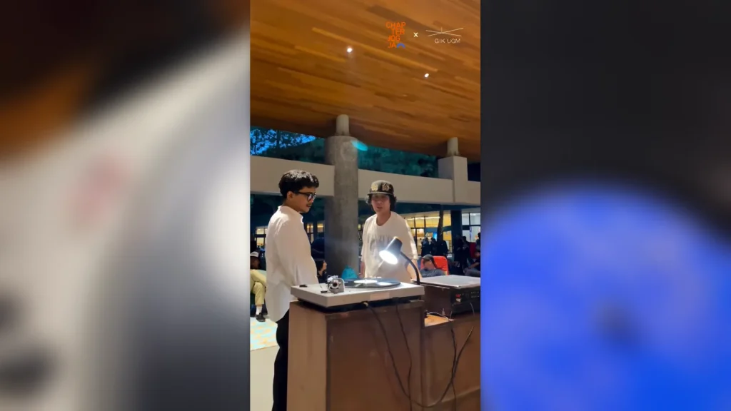 Dua pemuda berdiri di belakang meja DJ dengan pemutar piringan hitam di acara Penutupan Chapter Jogja x GIK UGM, dikelilingi pengunjung di area Joglo.