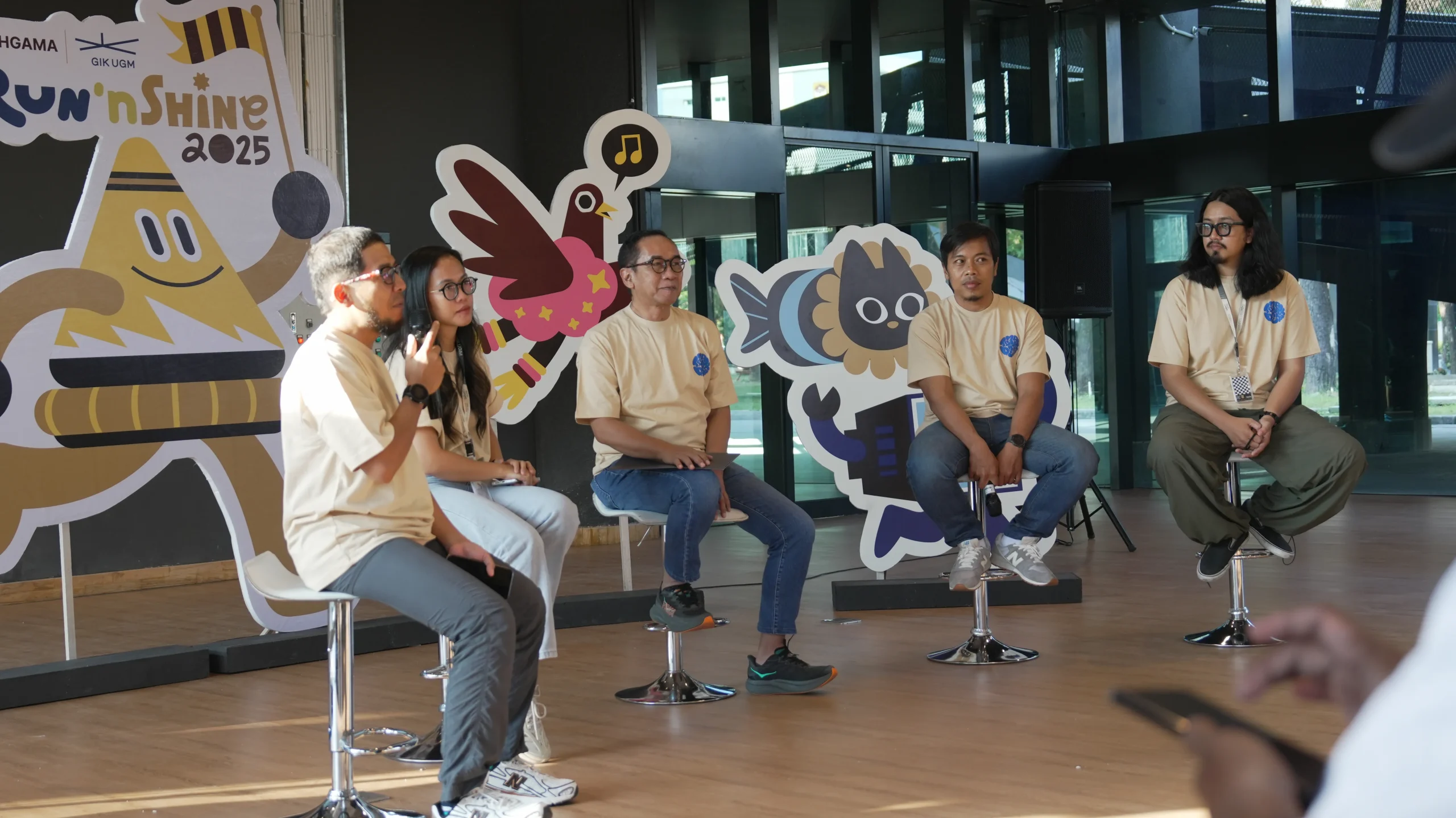 Empat orang duduk di atas kursi bar menghadap audiens, berbicara tentang acara Jogja Run’nShine 2025, dengan latar belakang dekorasi maskot acara yang unik.