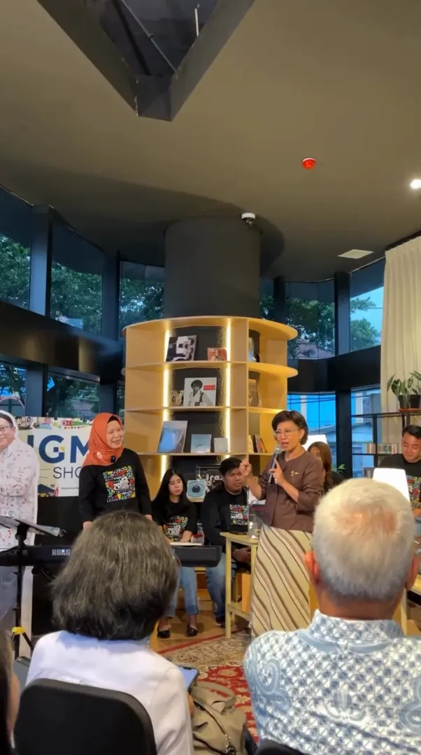 Prof. Adi Utarini berdiri berbicara di hadapan audiens dalam acara menuju Konser Amal, dikelilingi para musisi dan peserta dalam suasana hangat dan penuh inspirasi.
